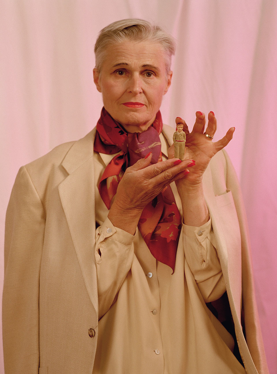 Image from The Cosmic Dominatrix. Linn as Dominatrix of Curators in white suit with red scarf. Linn is holding a small toy figure of a man.