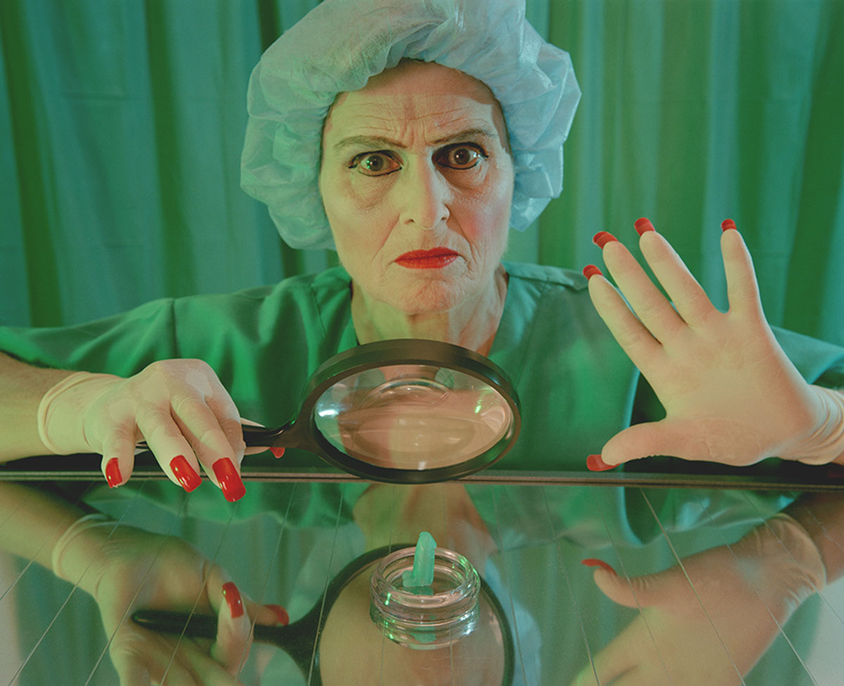 The Dominatrix of Gender from the series The Cosmic Dominatrix. She is wearing hospital scrubs and holding a magnifying glass. In front of her is a tiny toy penis in a specimen dish.