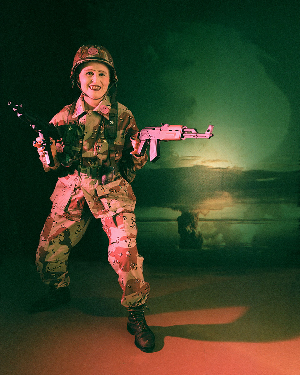 The Dominatrix of War from the series The Cosmic Dominatrix. She is wearing camouflage and carrying two machine guns.